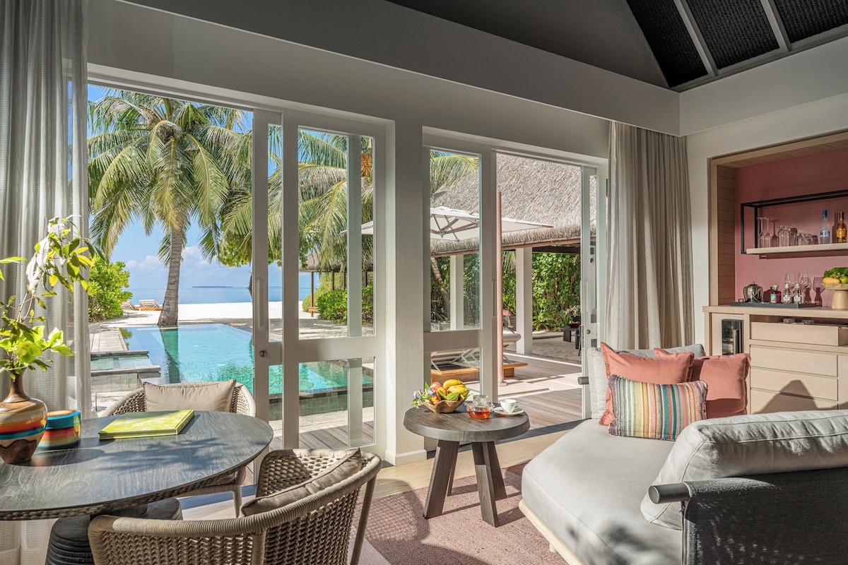 Una habitación con vista a una piscina y palmeras, un comedor con mesa y sillas, y una zona de estar con sofá y cojines.