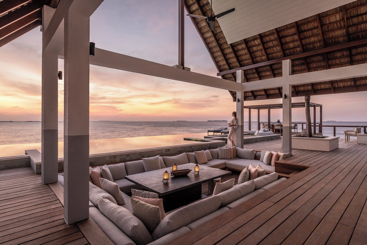 Amplia zona de estar al aire libre con vistas al océano al atardecer, que cuenta con una terraza de madera, asientos acolchados y una mesa con velas.
