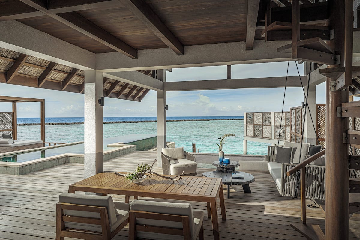 Eine Terrasse im Freien mit Holztisch und Stühlen, einer Sitzecke und einem kleinen runden Tisch mit einer Vase. Die Terrasse bietet Blick auf ein Gewässer und den Ozean.