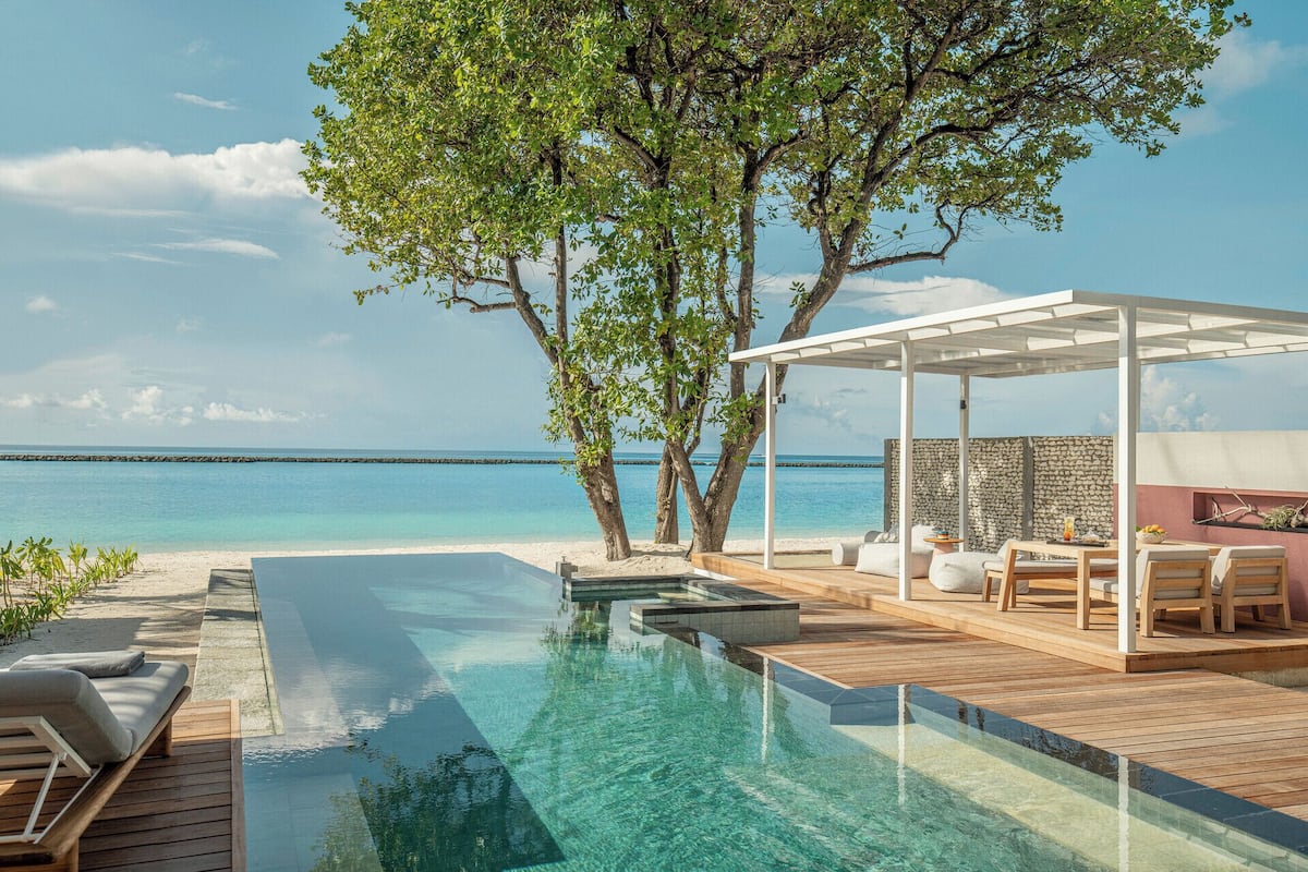Ein Infinity-Pool mit freiem Blick auf den Ozean, ein überdachter Sitzbereich mit Tischen und Stühlen sowie ein Baum, der Schatten spendet.