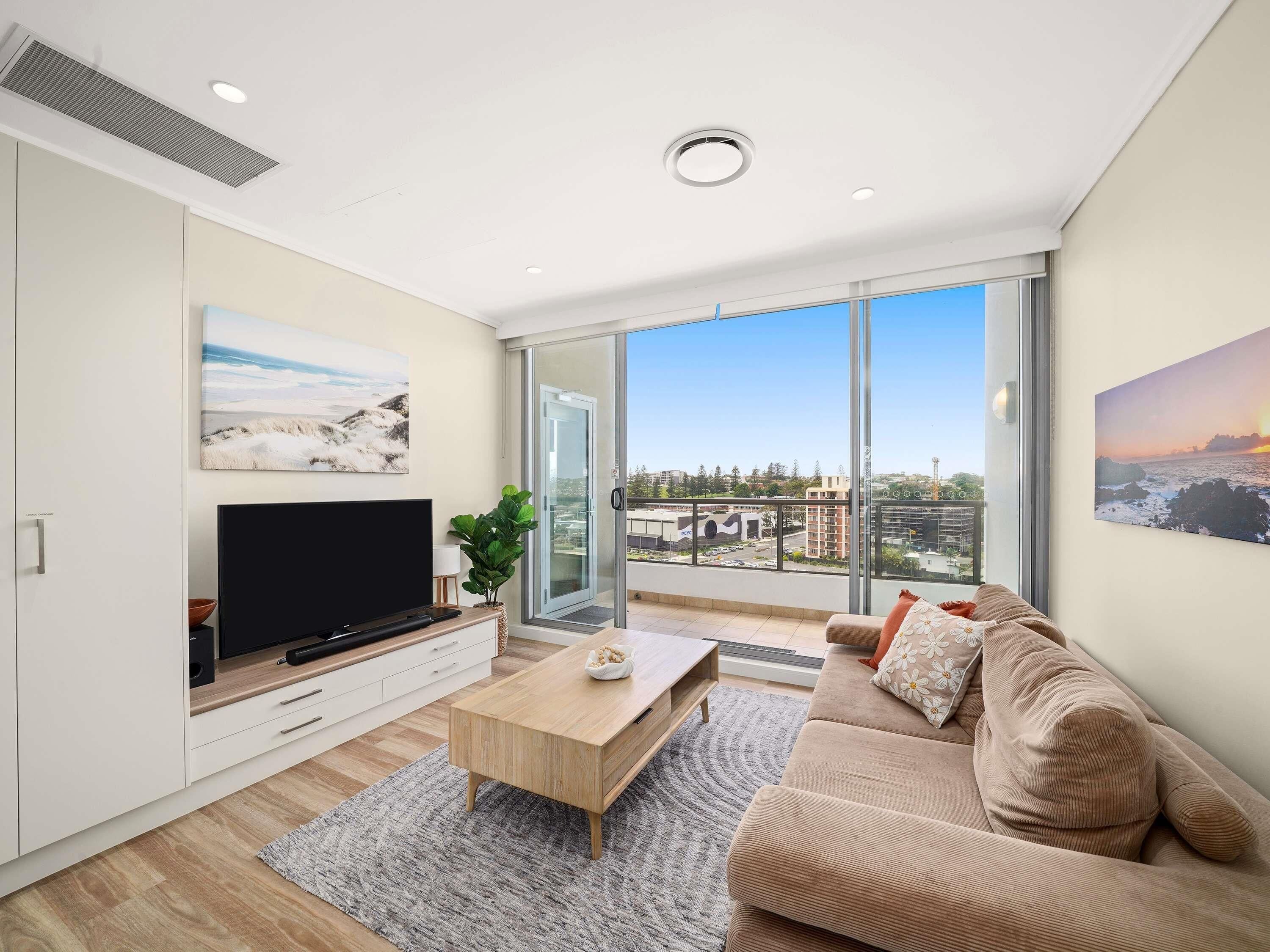 A modern living room with a flat-screen TV, a wooden coffee table, a beige sofa, and a large window offering a city view.