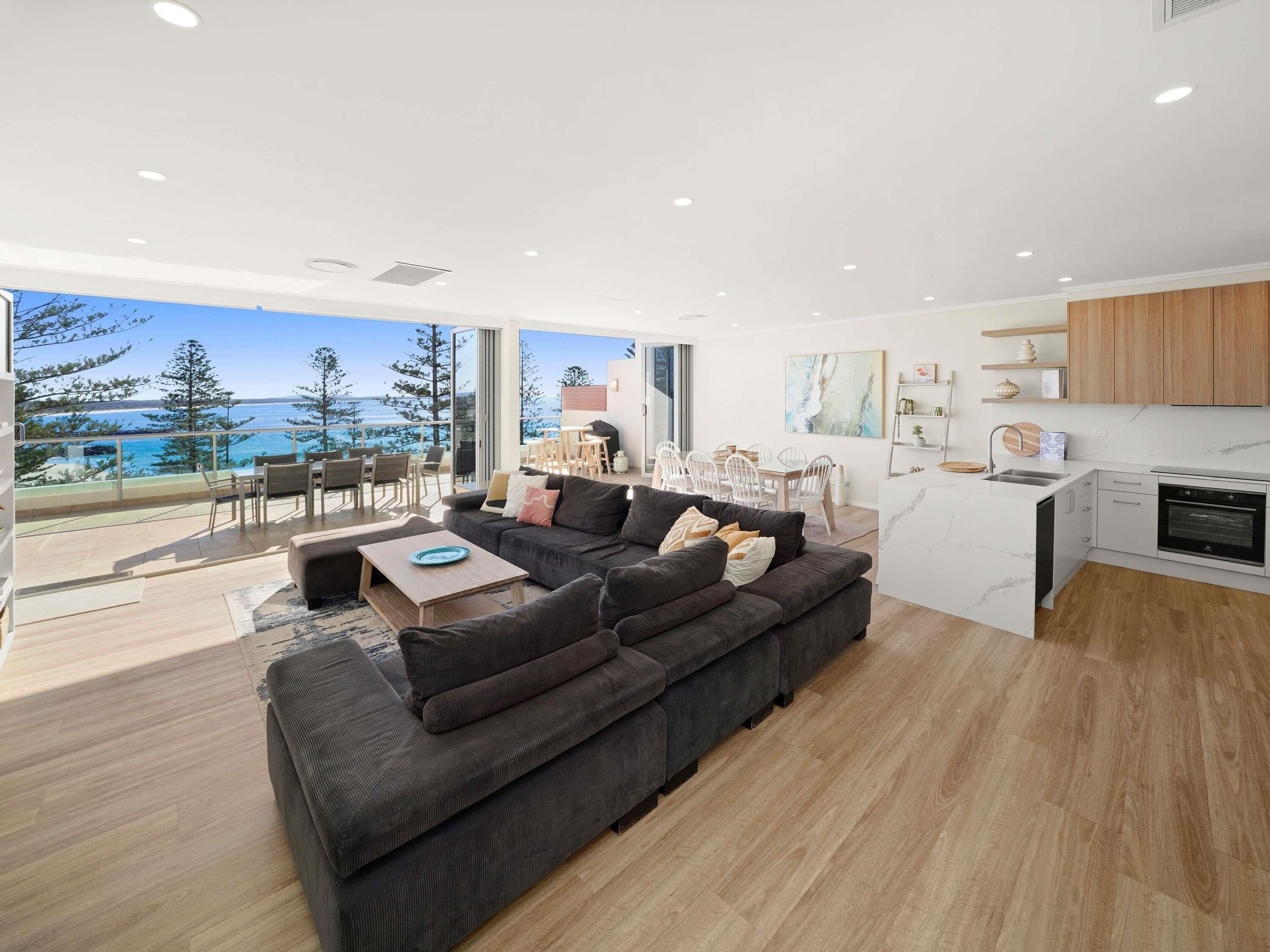 A modern living room with a large sofa, dining area, kitchen, and a balcony with a view of the sea.
