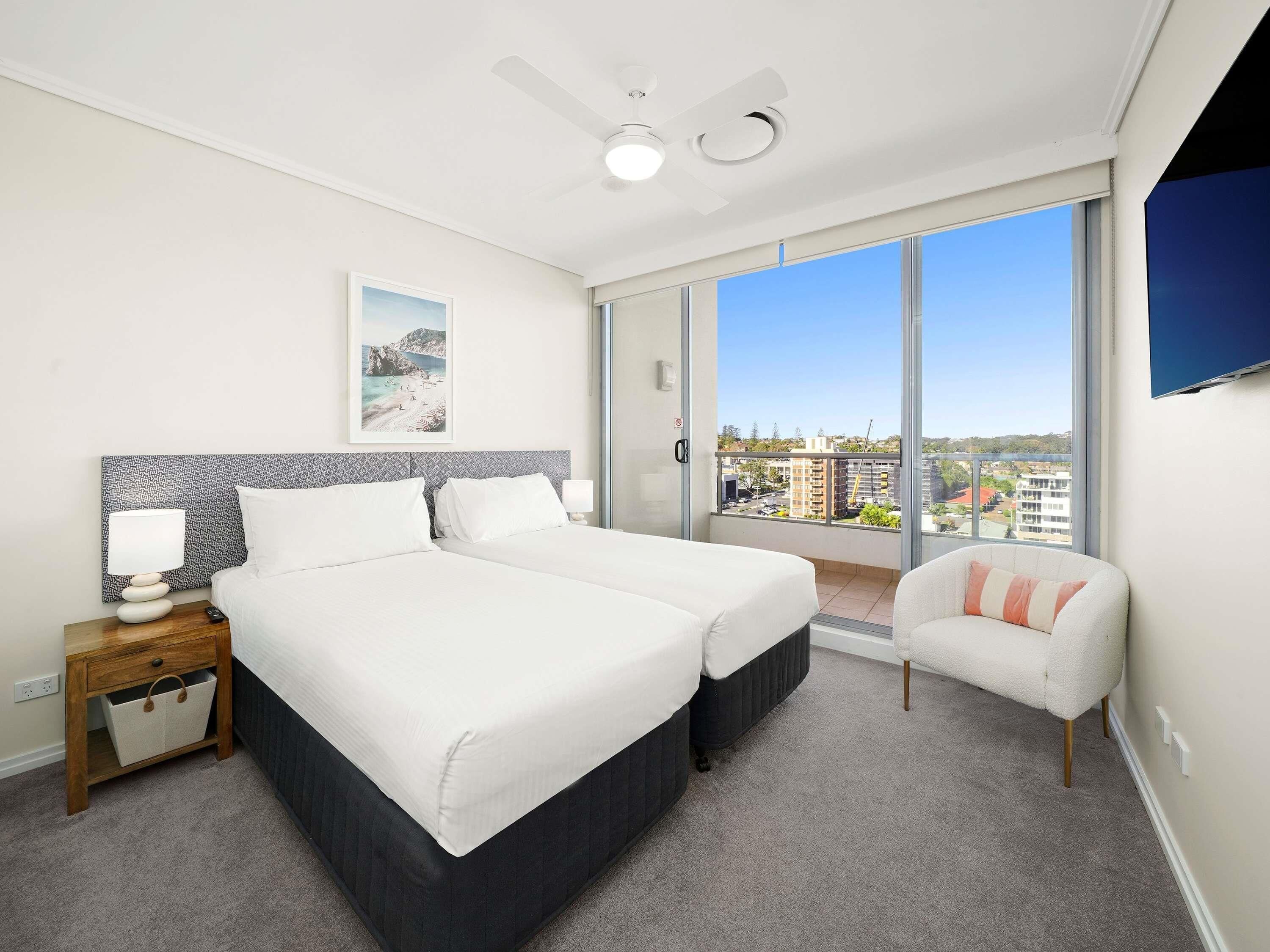 Ein Hotelzimmer mit Bett, Nachttisch, Sessel, Deckenventilator und großem Fenster mit Blick auf die Stadt.