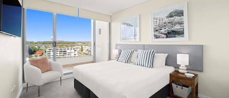 Un dormitorio moderno con una cama grande, una mesita de noche con lámpara, una silla y vistas a la ciudad a través de amplias ventanas.