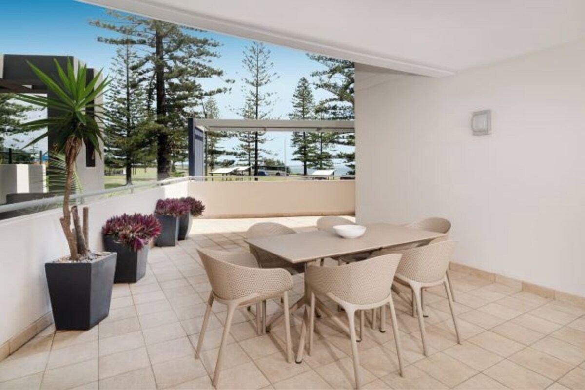 Un área de comedor exterior moderna con una mesa, sillas, plantas en macetas y vistas a árboles y una masa de agua.