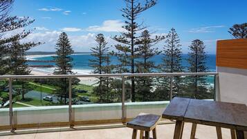 A balcony with a view of the ocean and beach, a wooden table, and a railing.