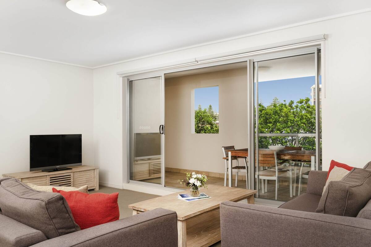 A modern living room with a flat-screen TV, grey sofas, a wooden coffee table, and a sliding glass door leading to an outdoor balcony.