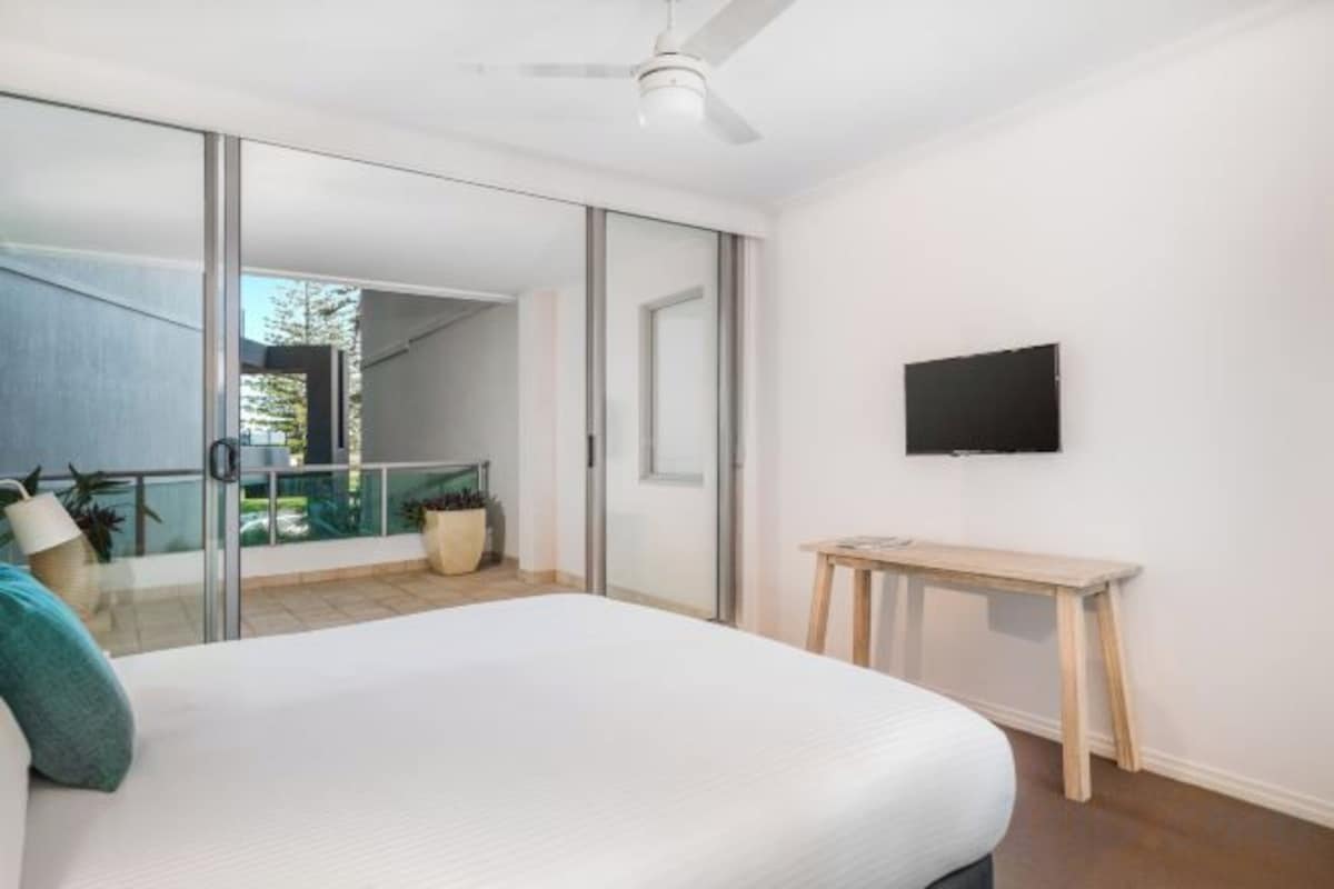 Un dormitorio con cama, televisor, puerta corrediza de vidrio y balcón con plantas.