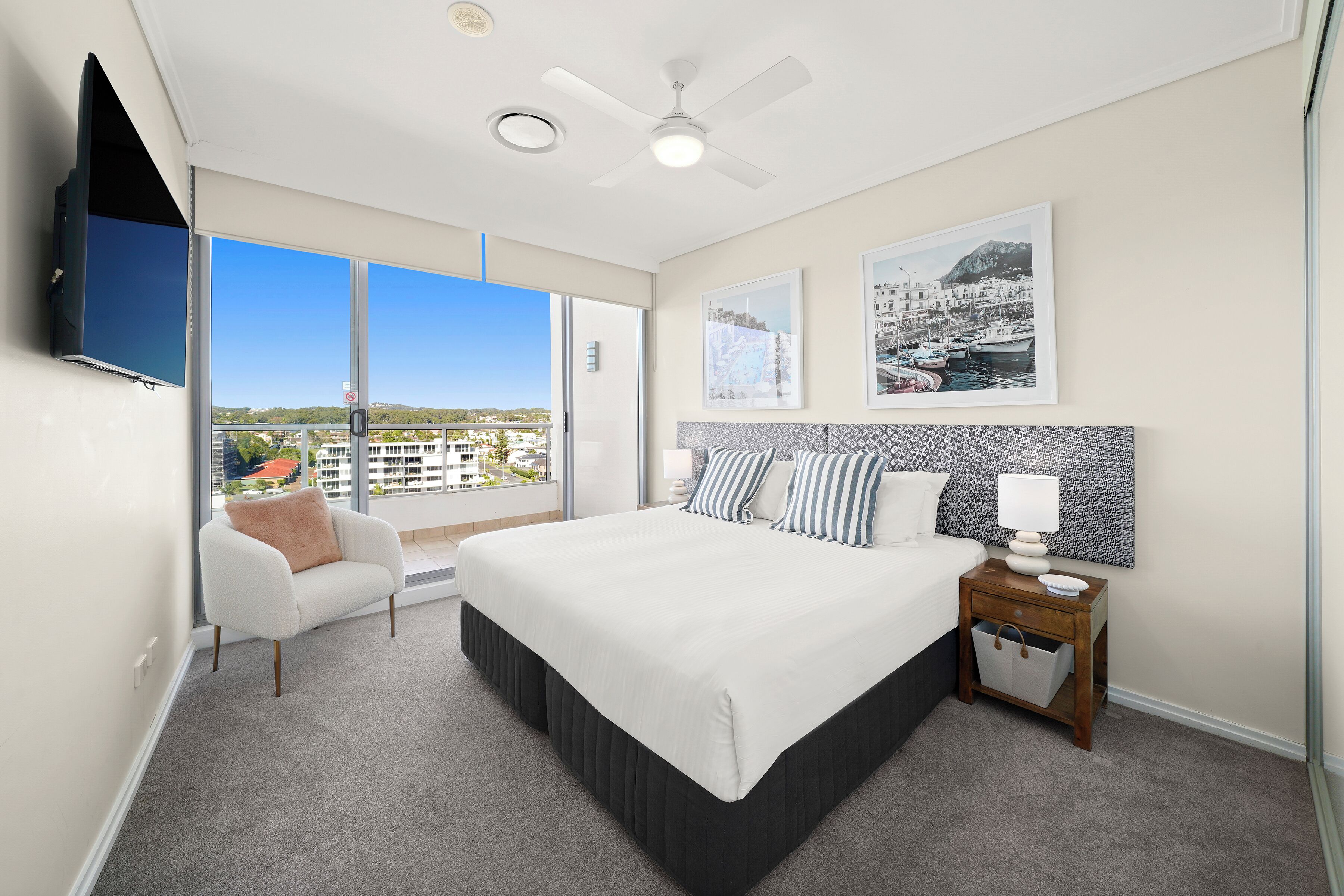 Een moderne slaapkamer met een groot bed, een aan de muur bevestigde televisie, een plafondventilator en een balkon met uitzicht over de stad.