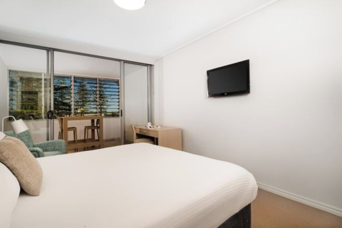 A bedroom with a bed, a desk, a chair, a TV, and a sliding glass door leading to a balcony.