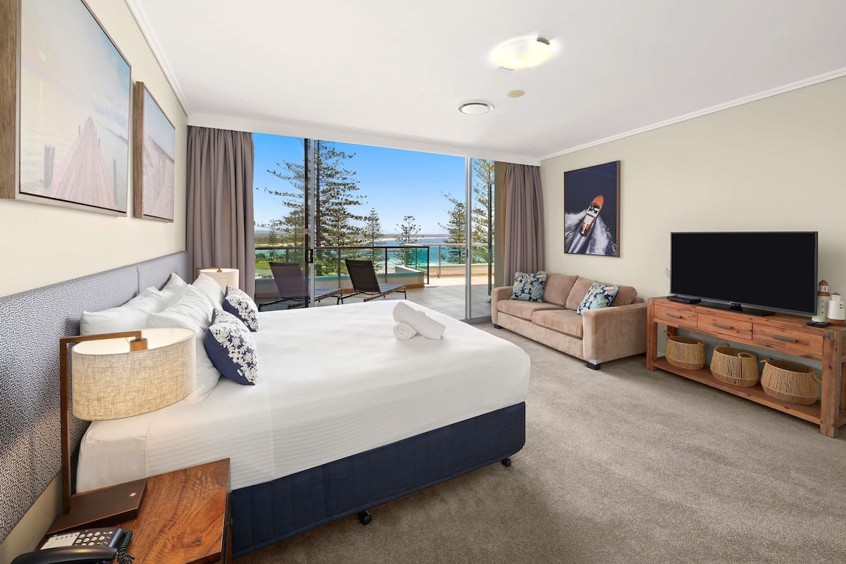A hotel room with a large bed, a sofa, a TV, and a balcony with a view.