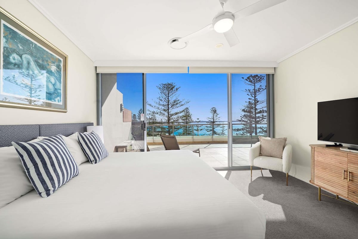 A modern hotel room with a large bed, a ceiling fan, a TV, a balcony with a view of trees and the sea, and a painting on the wall.