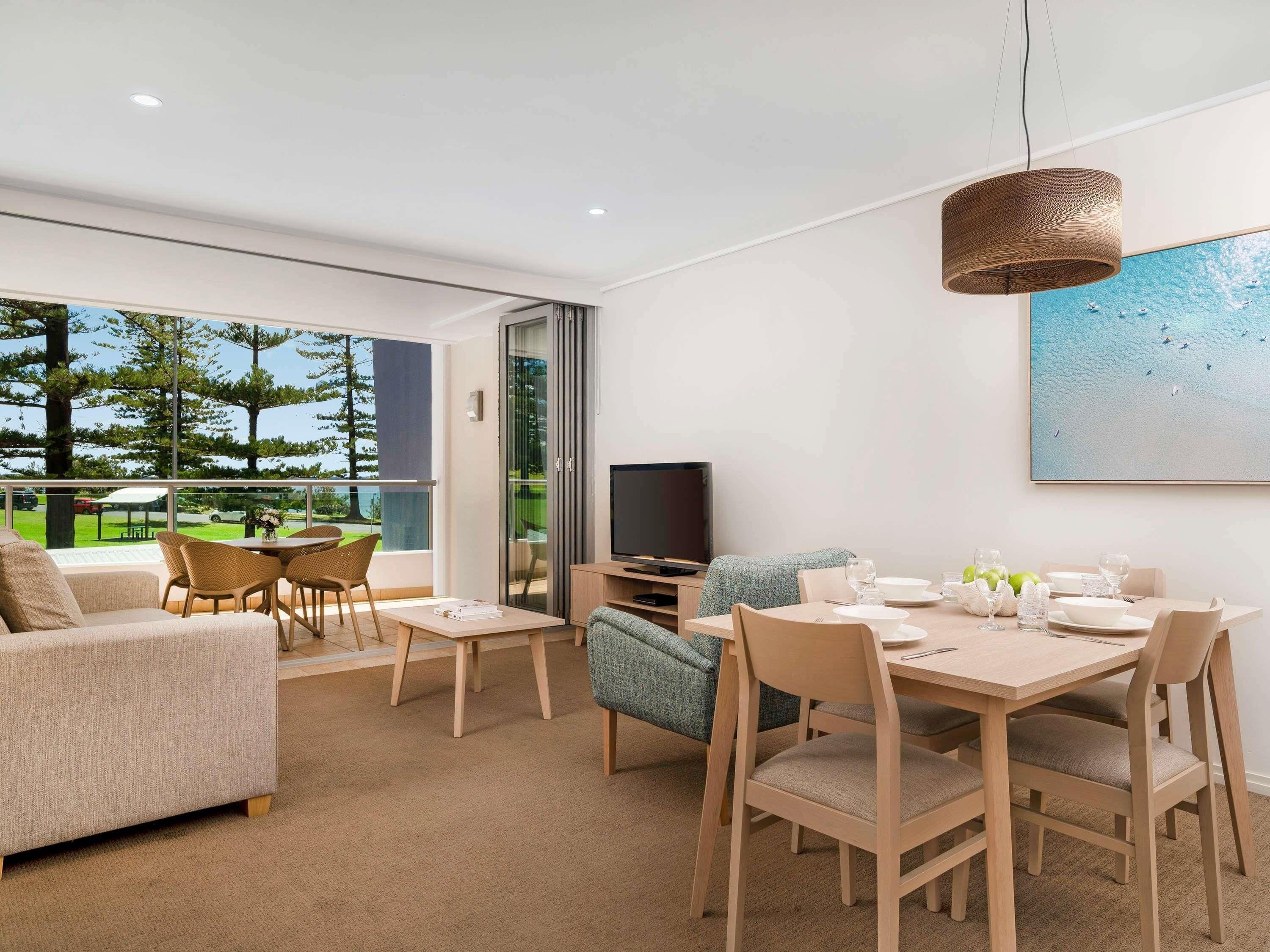A modern living room with a dining area, large windows, a flat-screen TV, and a view of the outdoors.