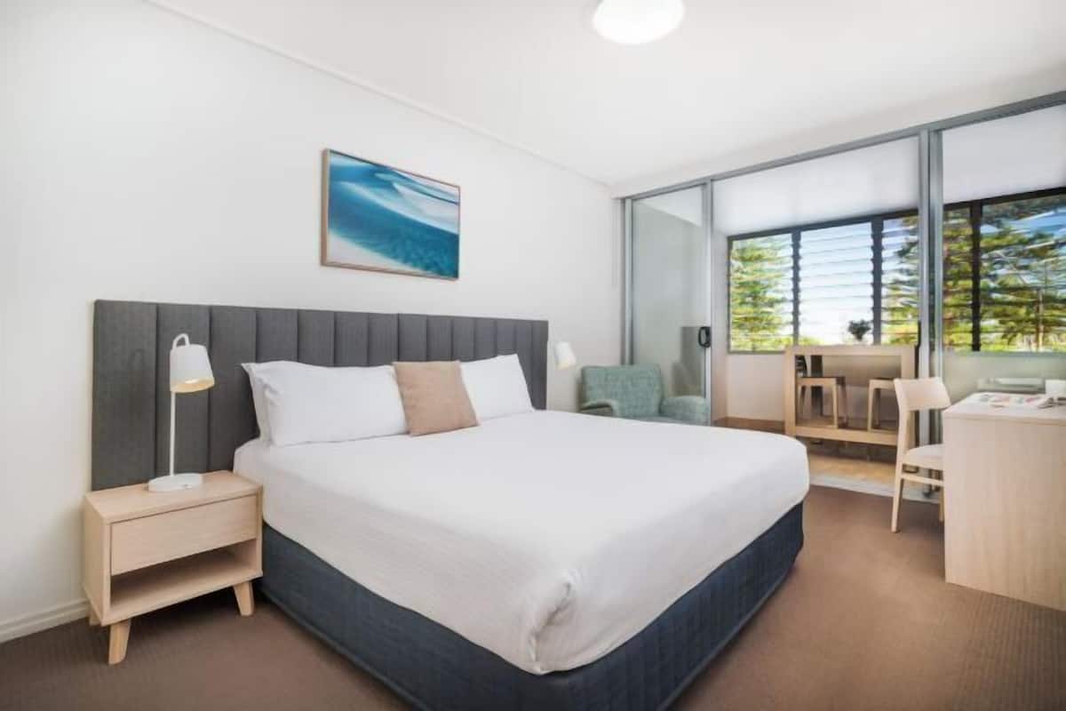 A modern hotel room with a bed, bedside table, lamp, and a view of the outdoors through a large window.