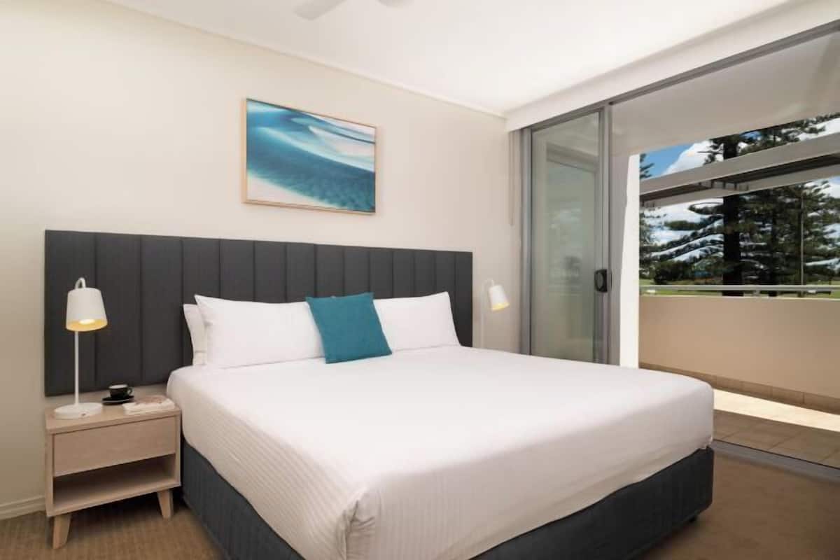 A modern hotel room with a large bed, a nightstand, a bedside lamp, a window with a view, and a painting on the wall.