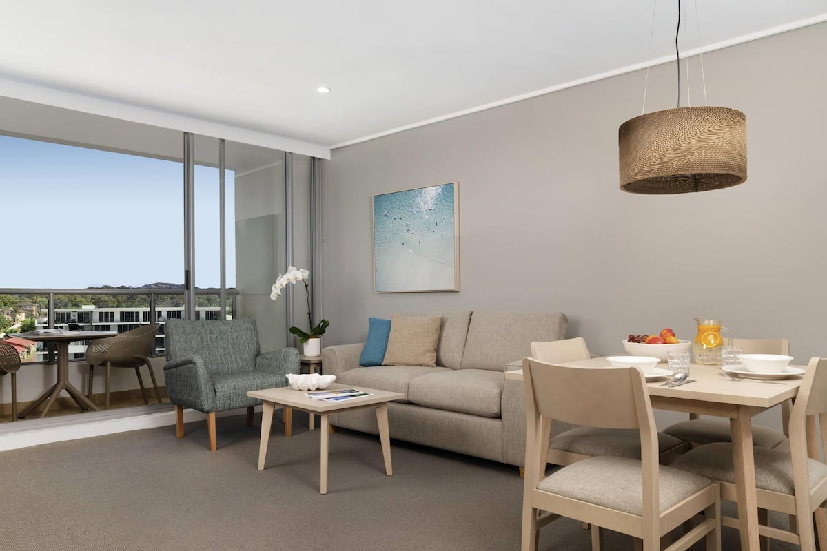 A modern living room with a sofa, chairs, a dining table, and a large window offering a view.