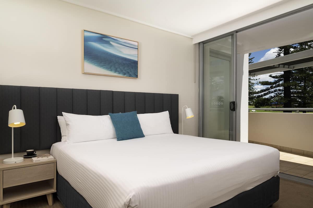 A modern hotel room with a large bed, a nightstand, a bedside lamp, a framed artwork, and a glass door leading to a balcony.