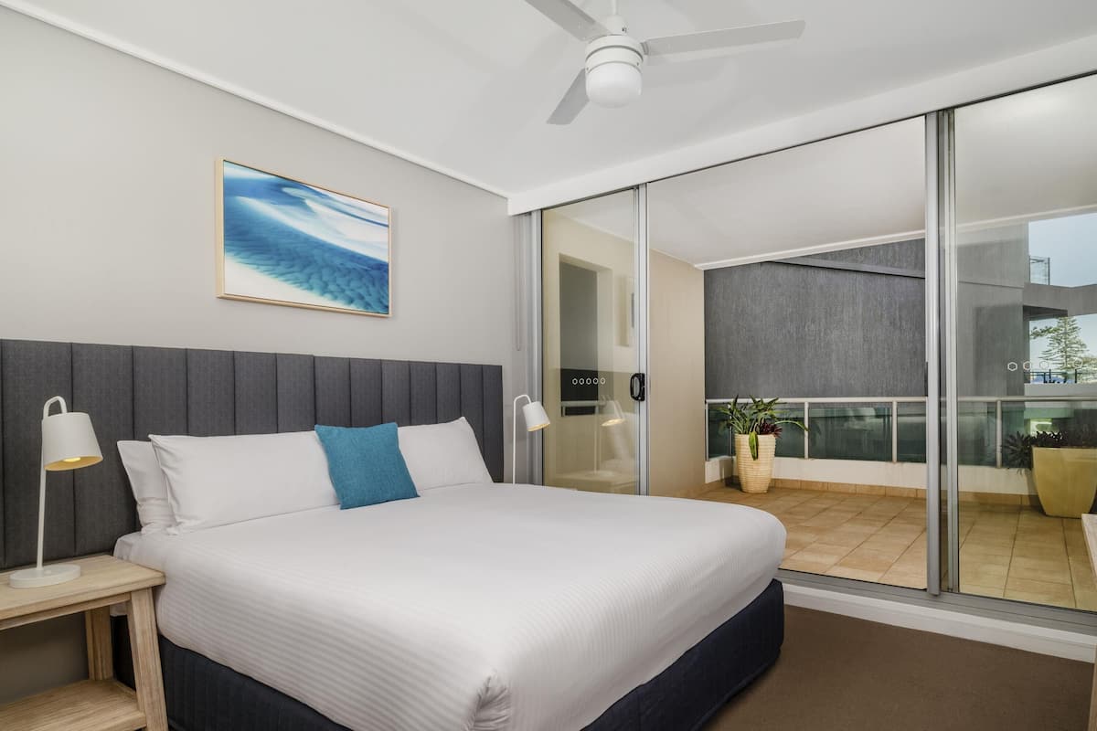 A modern hotel room with a large bed, a ceiling fan, a view through a sliding glass door, and a painting on the wall.