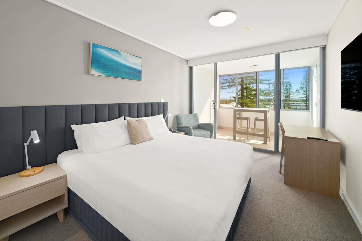 A modern hotel room with a large bed, a desk, a chair, and a view of the outdoors.