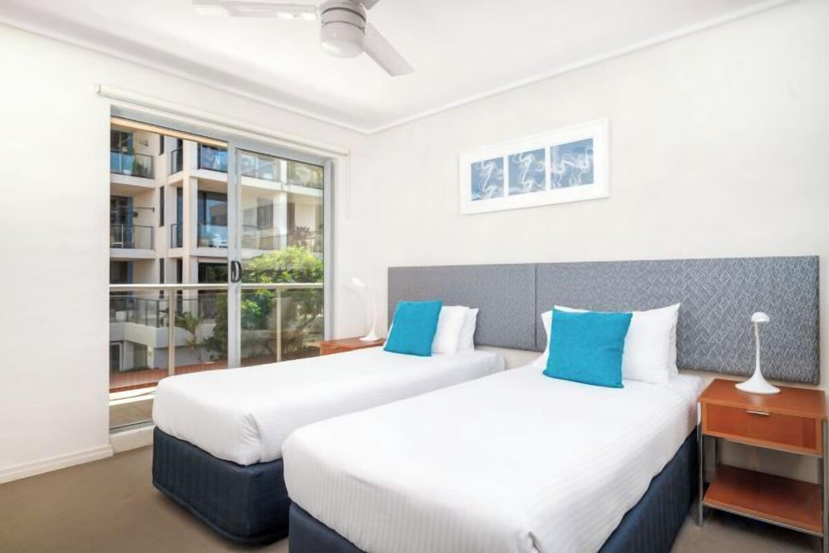 A hotel room with two beds, a window with a view, a ceiling fan, and a framed picture on the wall.
