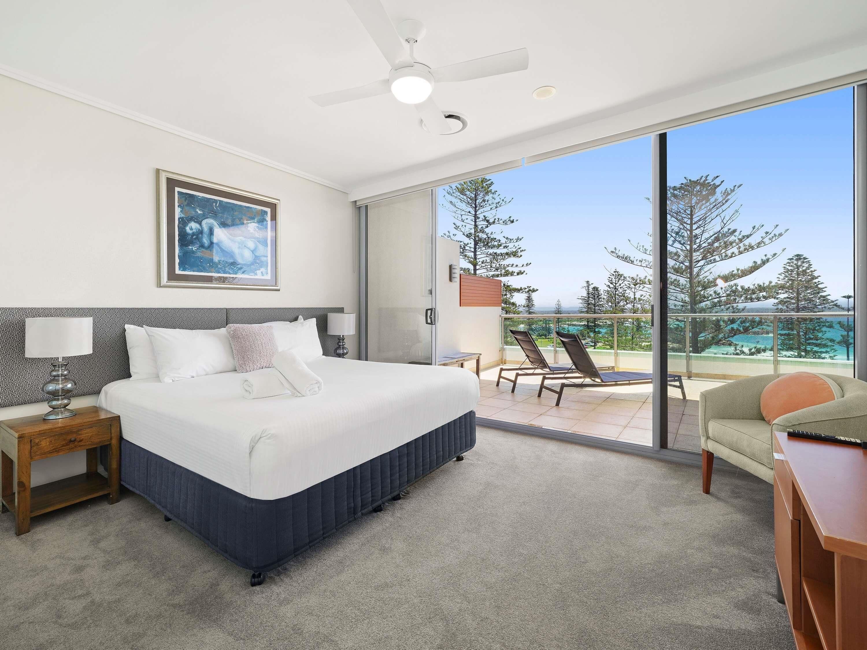 A hotel room with a bed, a nightstand, a desk, a chair, a balcony with a view, and a ceiling fan.
