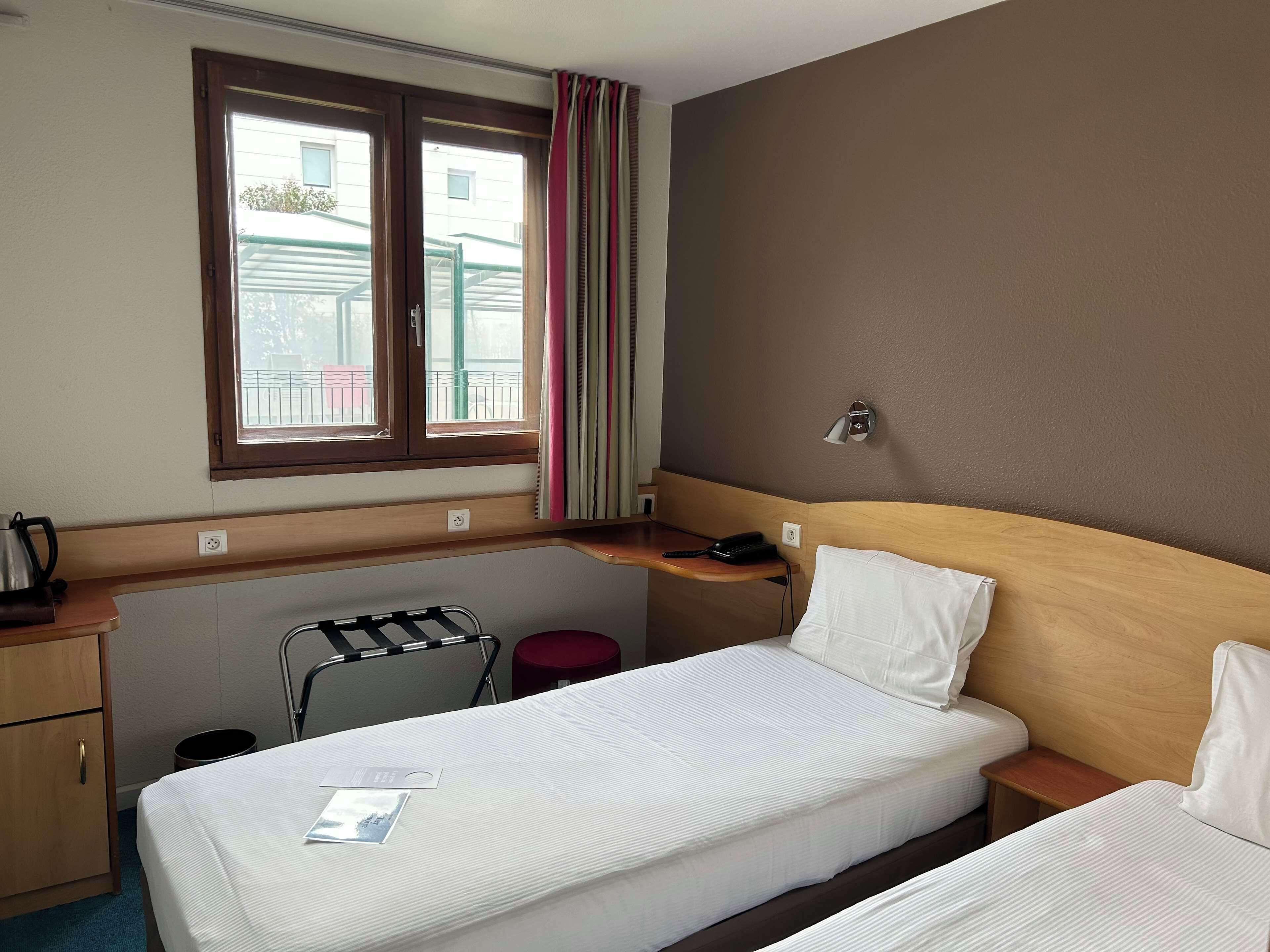Quarto de hotel com cama, escrivaninha, cadeira, mesinha e janela com cortinas.