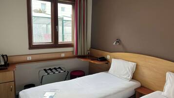 Quarto de hotel com cama, escrivaninha, cadeira, mesinha e janela com cortinas.