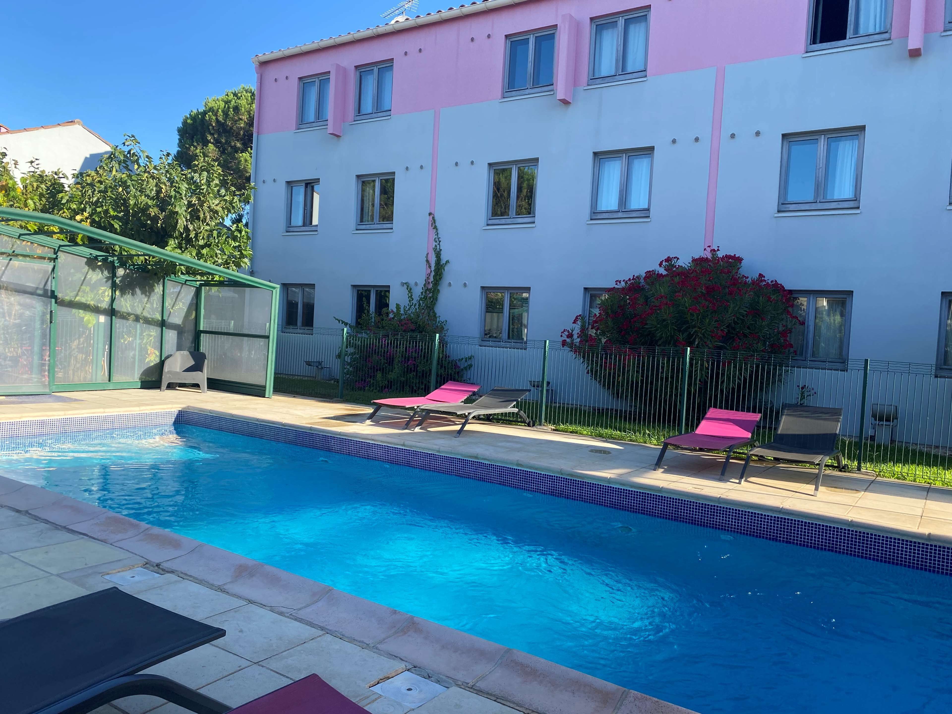 A swimming pool with lounge chairs, a covered patio, and a building with pink and white exterior walls.