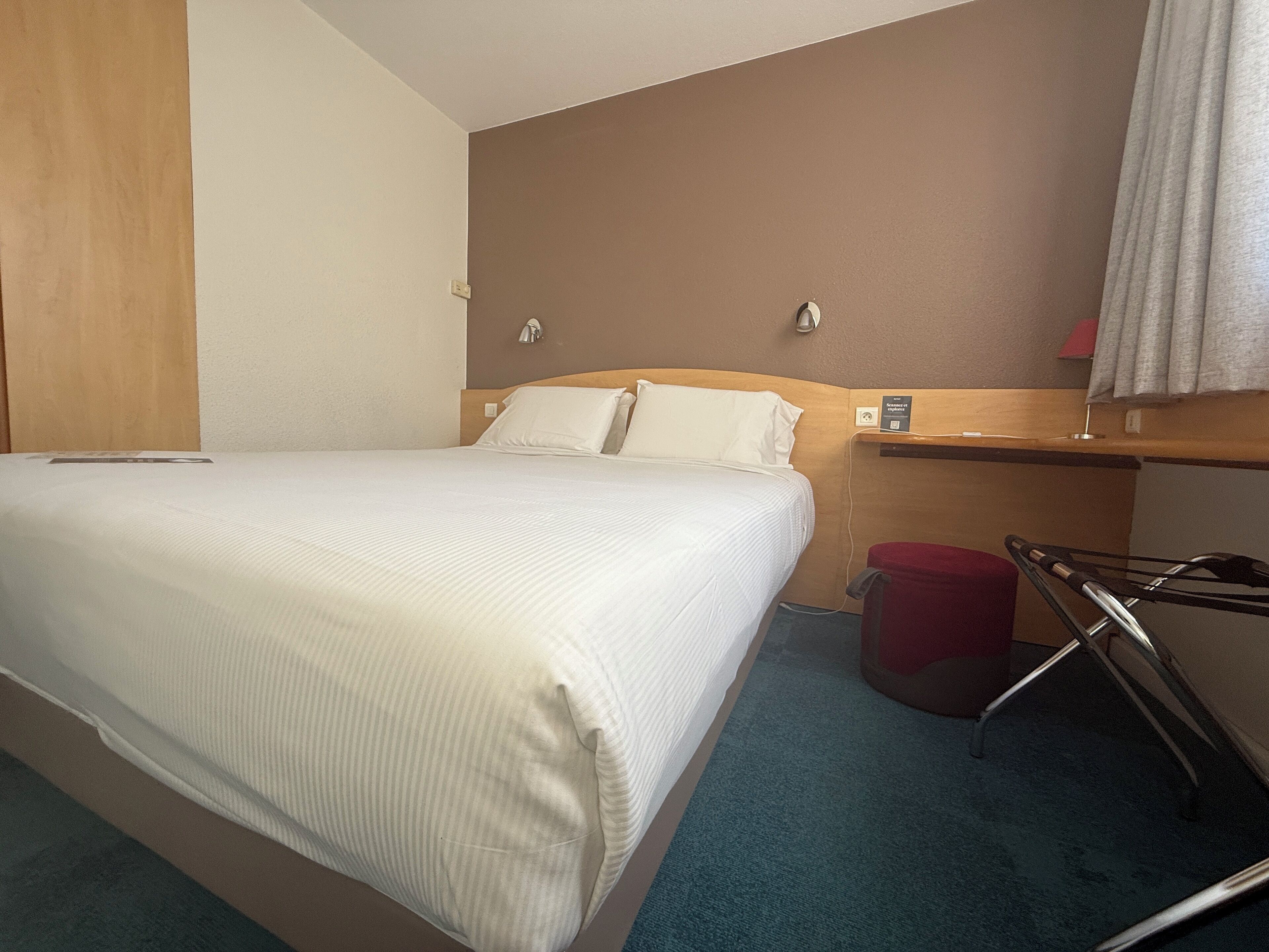 A hotel room with a bed, a wooden headboard, a red trash bin, and a wall-mounted charging station.