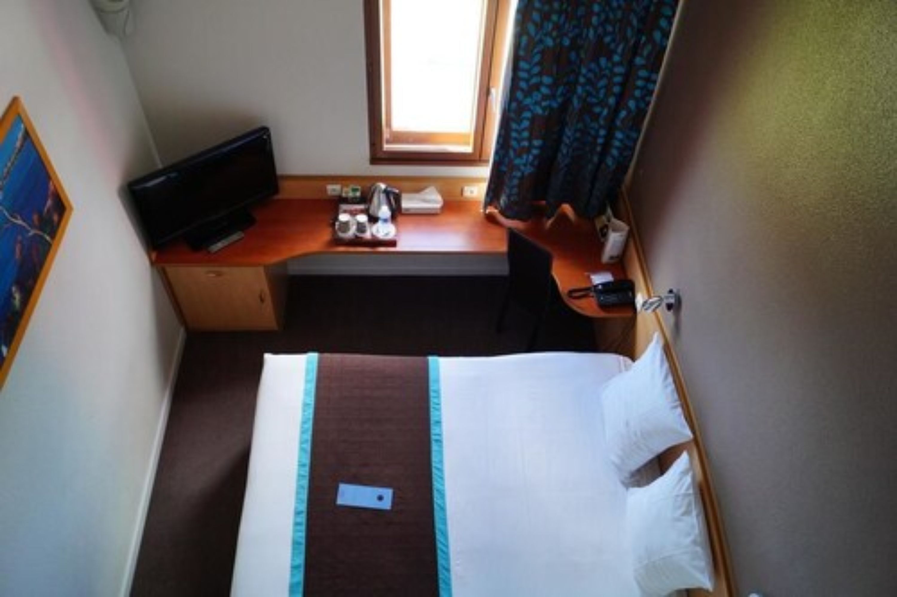 Ett litet hotellrum med en säng, ett skrivbord med en dator, ett fönster och en gardin.