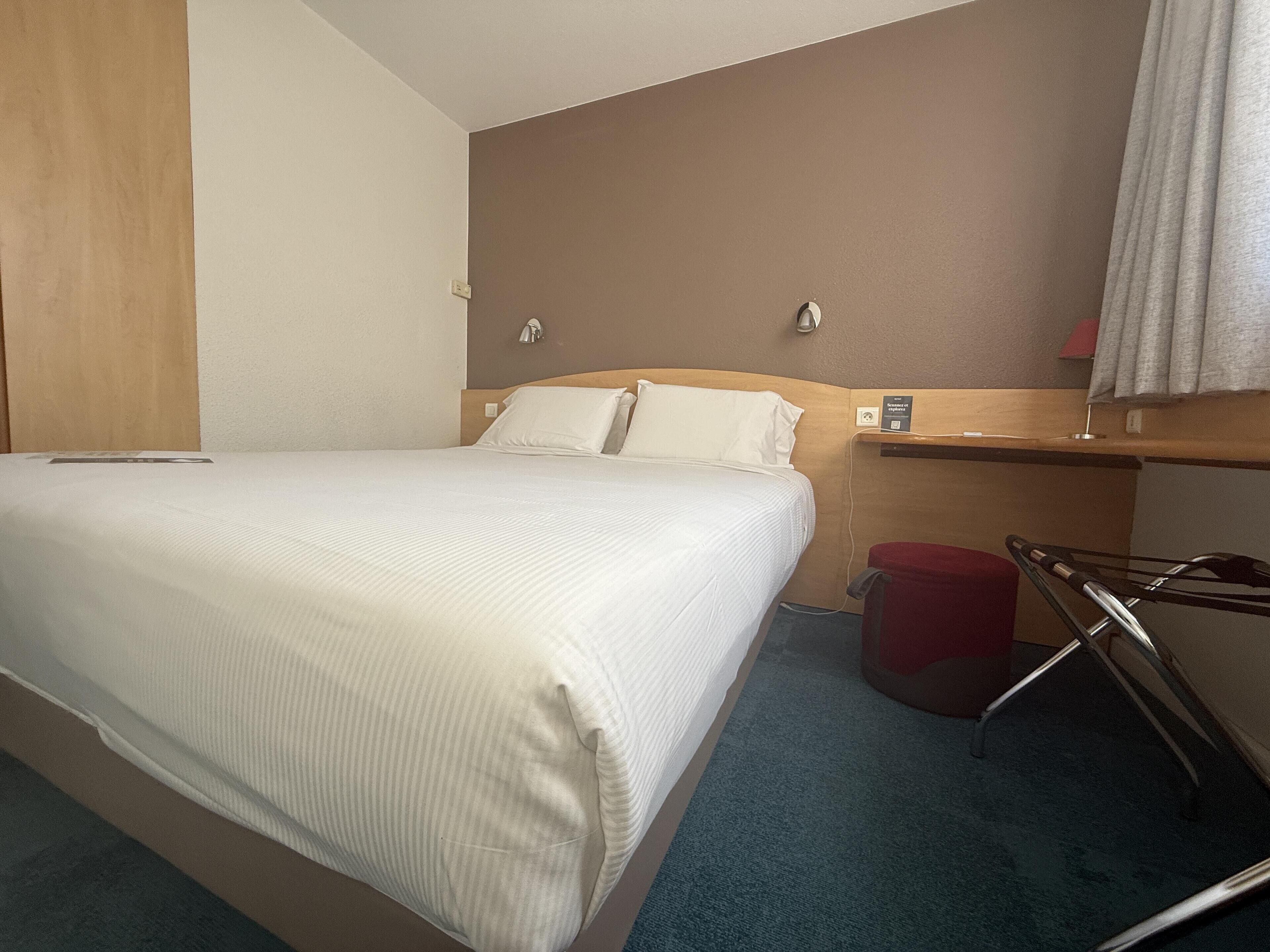 Habitación de hotel con cama, cabecera de madera, un cubo de basura rojo y una repisa de pared.