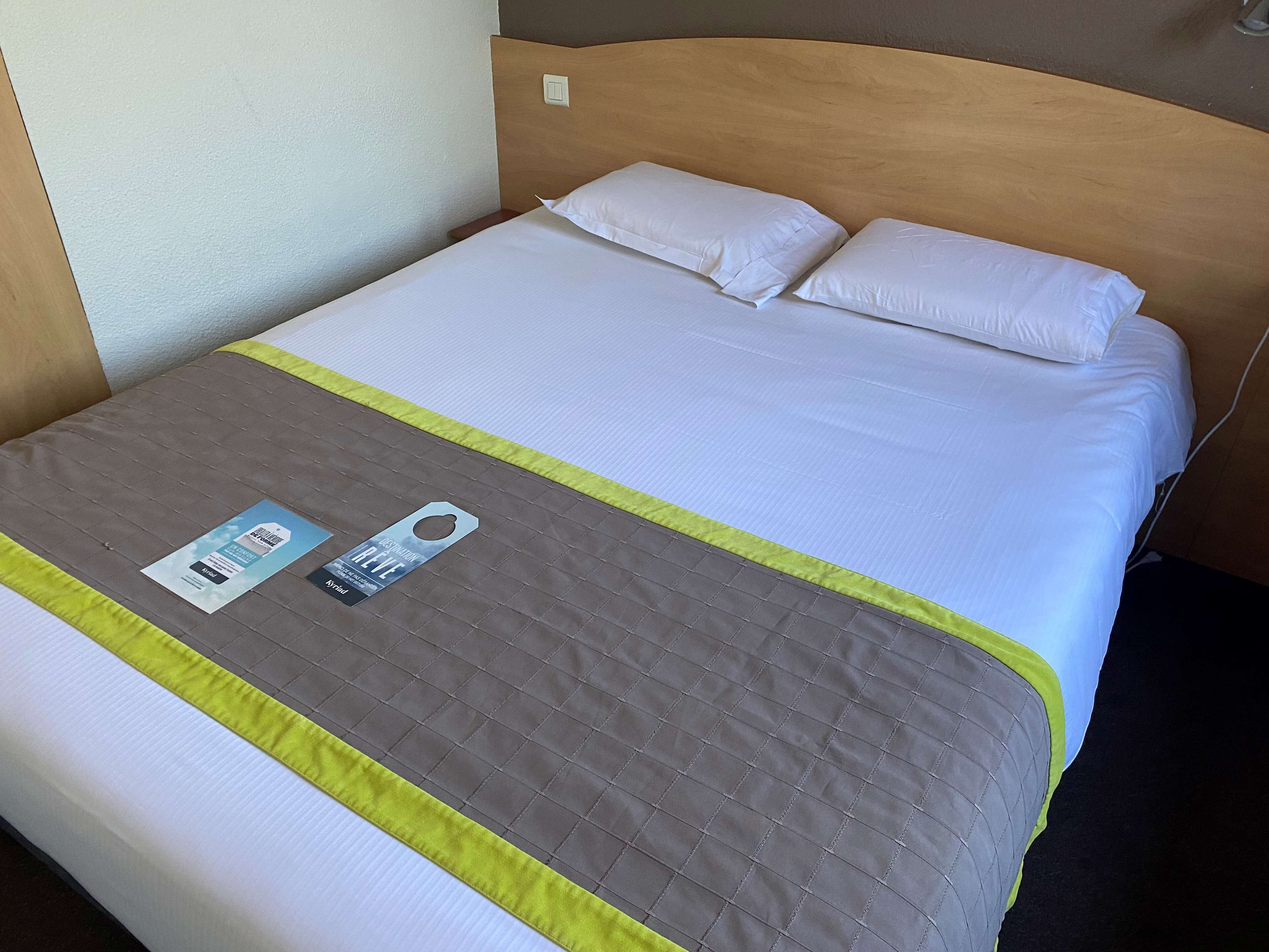 Una cama bien hecha con colcha gris y ribete amarillo, cabecera de madera y almohada blanca.