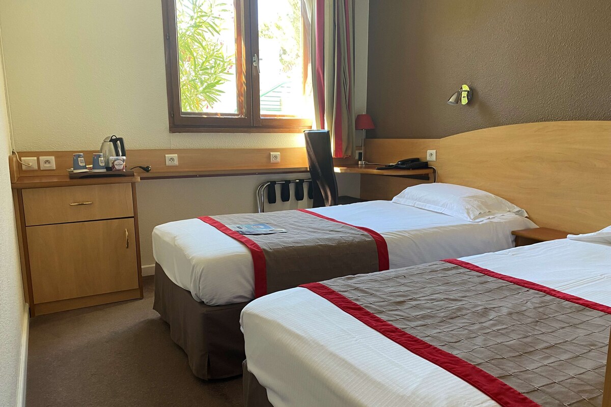 Habitación de hotel con dos camas, un escritorio, una silla y una ventana con vistas a zonas verdes.
