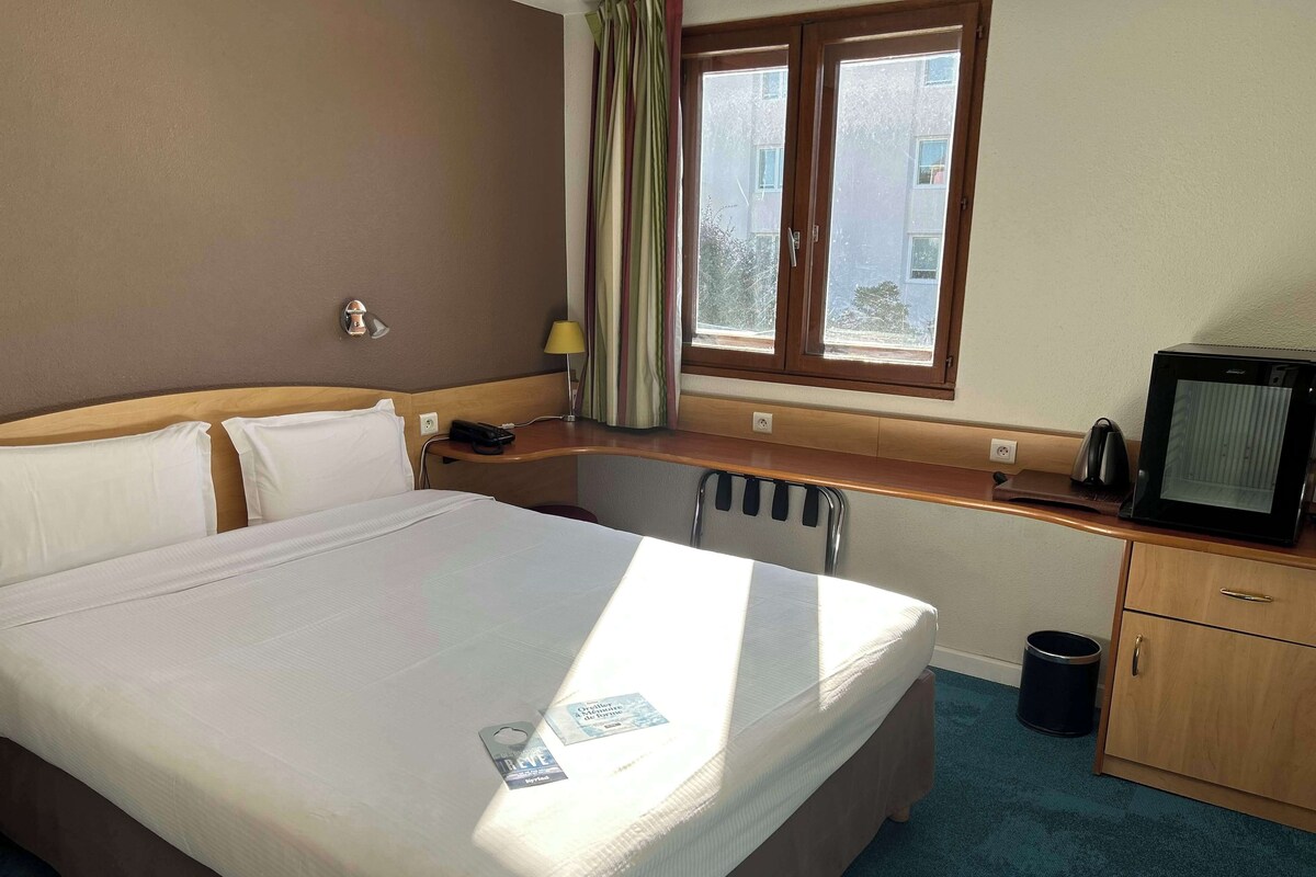 Habitación de hotel con una cama grande, un escritorio con computadora, televisión y ventana con cortinas.