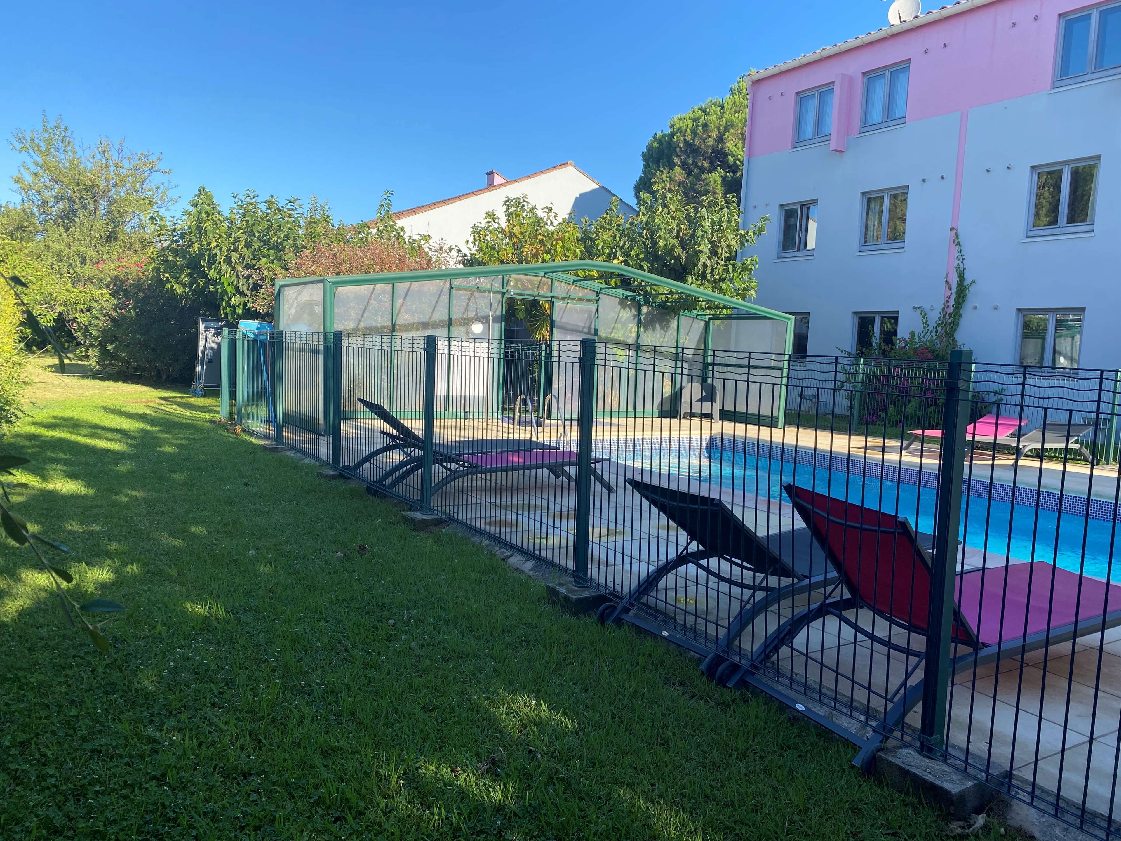 Una piscina con valla verde, zona cubierta y edificio con paredes exteriores en rosa y blanco.