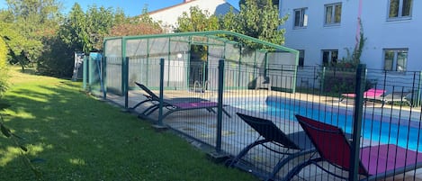 Una piscina con valla verde, zona cubierta y edificio con paredes exteriores en rosa y blanco.