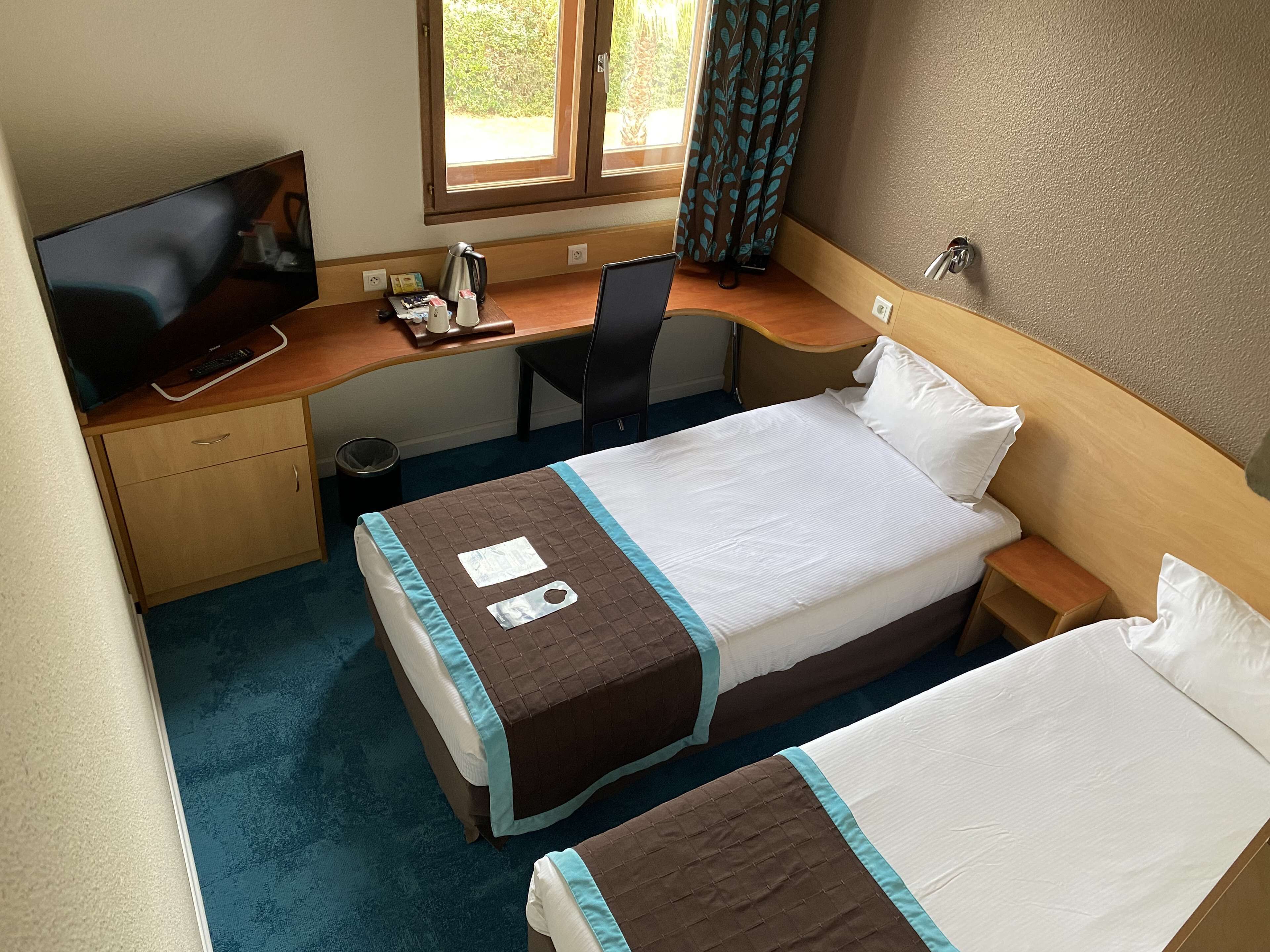 Et hotelværelse med to senge, et skrivebord med computer, et fjernsyn og et vindue med gardiner.