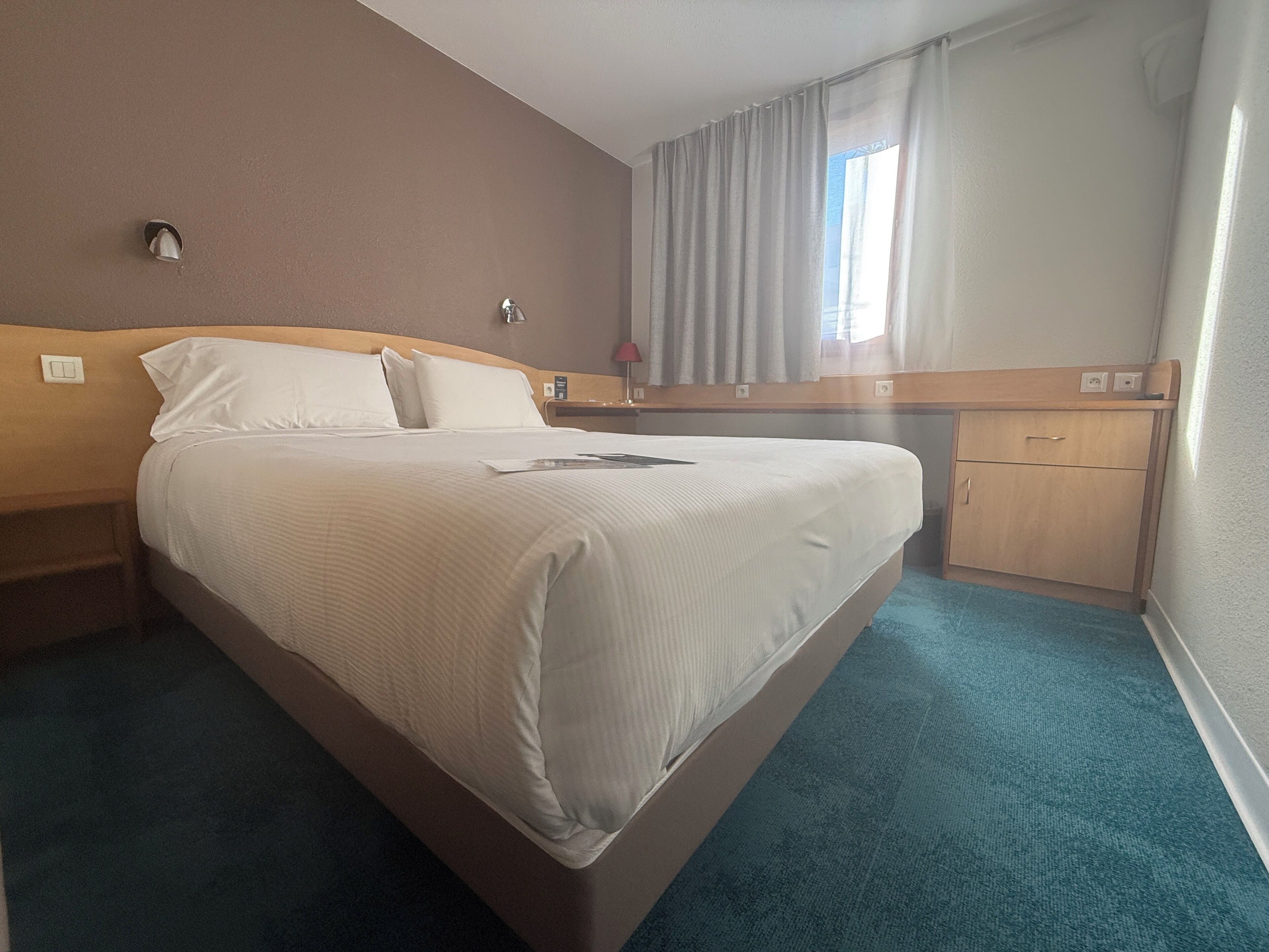 A hotel room with a large bed, a nightstand, a desk, and a window with curtains.