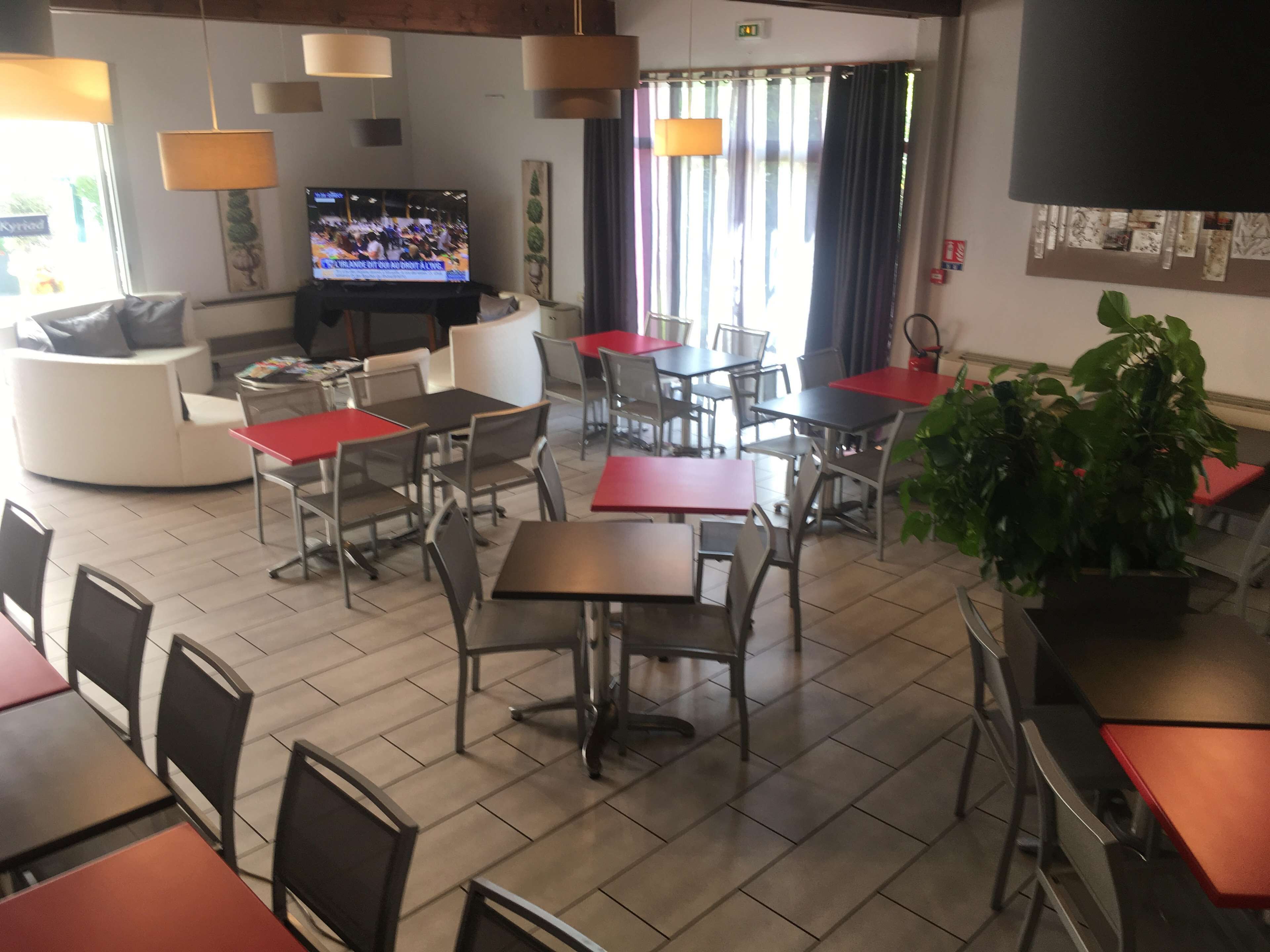 Een restaurant met meerdere tafels en stoelen, een televisie waarop een sportevenement te zien is, en een grote potplant.