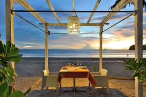 A beachside dining setup with a table and chairs under a pergola, overlooking the ocean.