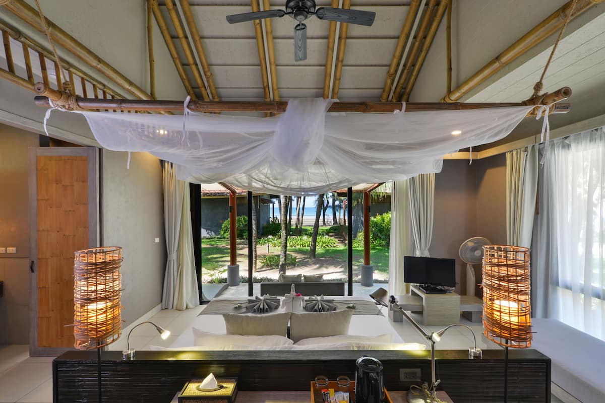 A modern hotel room with a large bed, a ceiling fan, a view of the beach, and two bedside lamps.