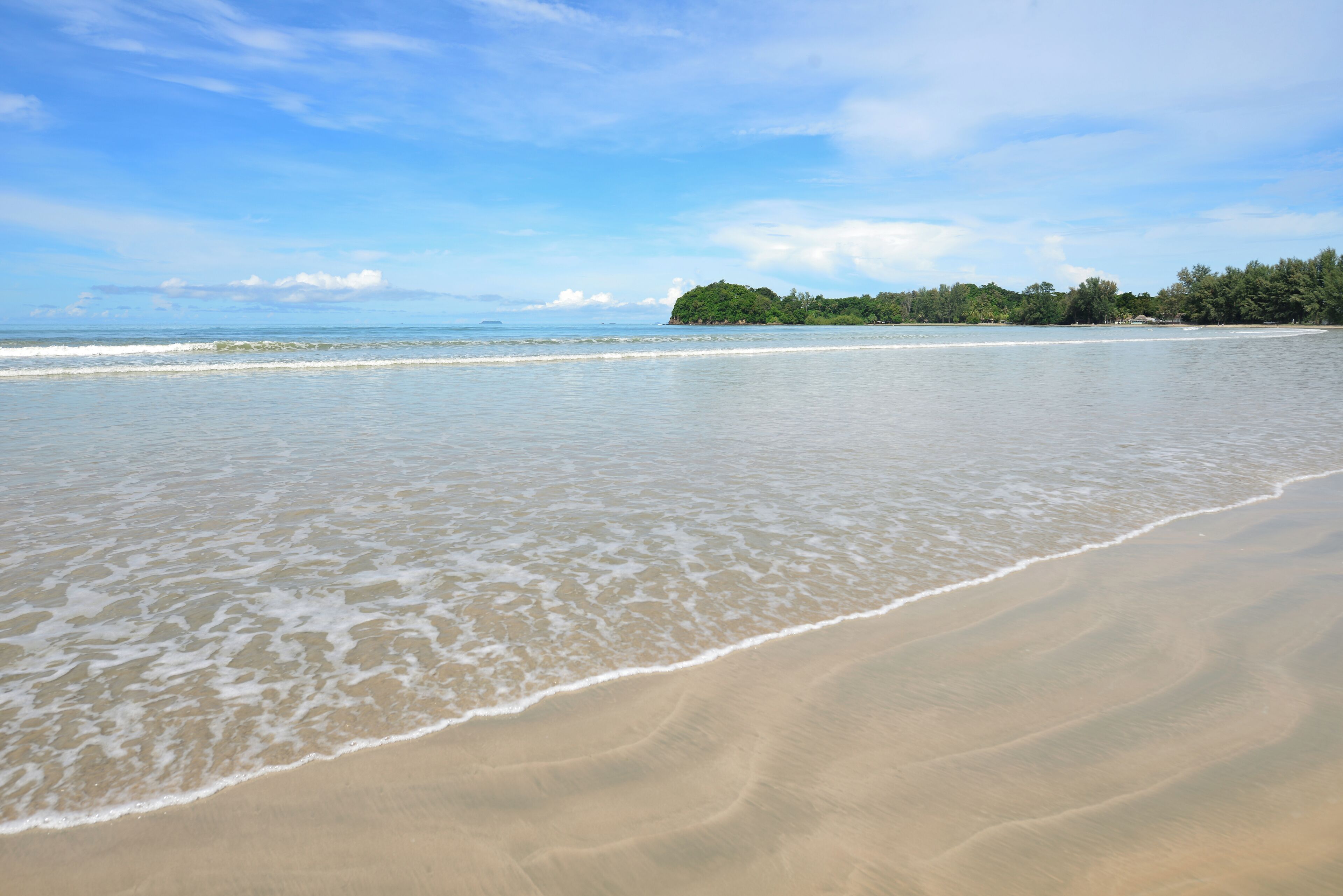 Ein Strand mit klarem Wasser, sanften Wellen und einer fernen, von Bäumen bewachsenen Küste.