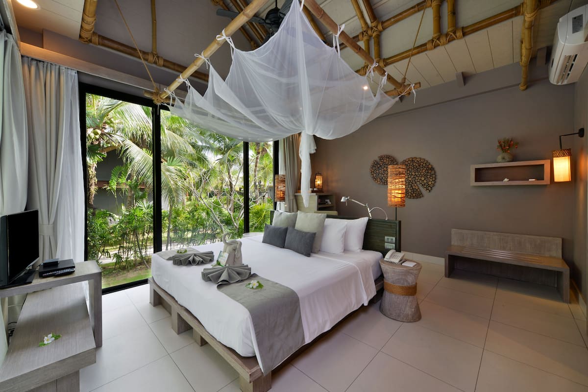 A modern bedroom with a large bed, a TV, a desk, and a view of lush greenery.