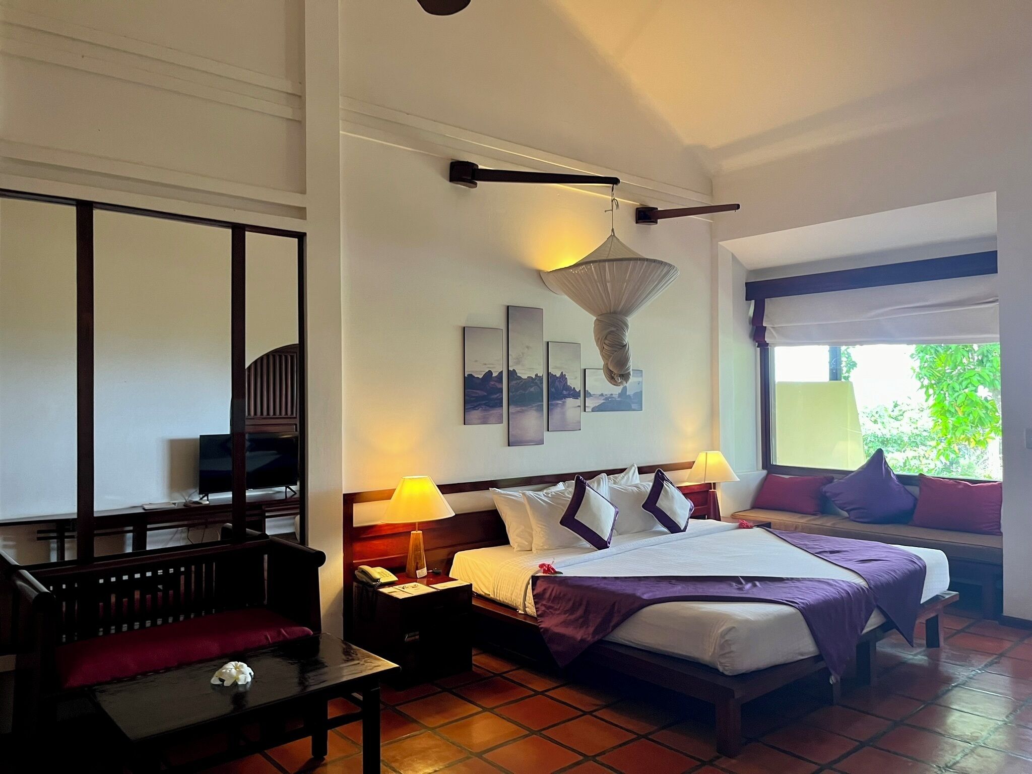 A hotel room with a bed, bedside tables, a desk, a chair, and a window with a view of greenery.