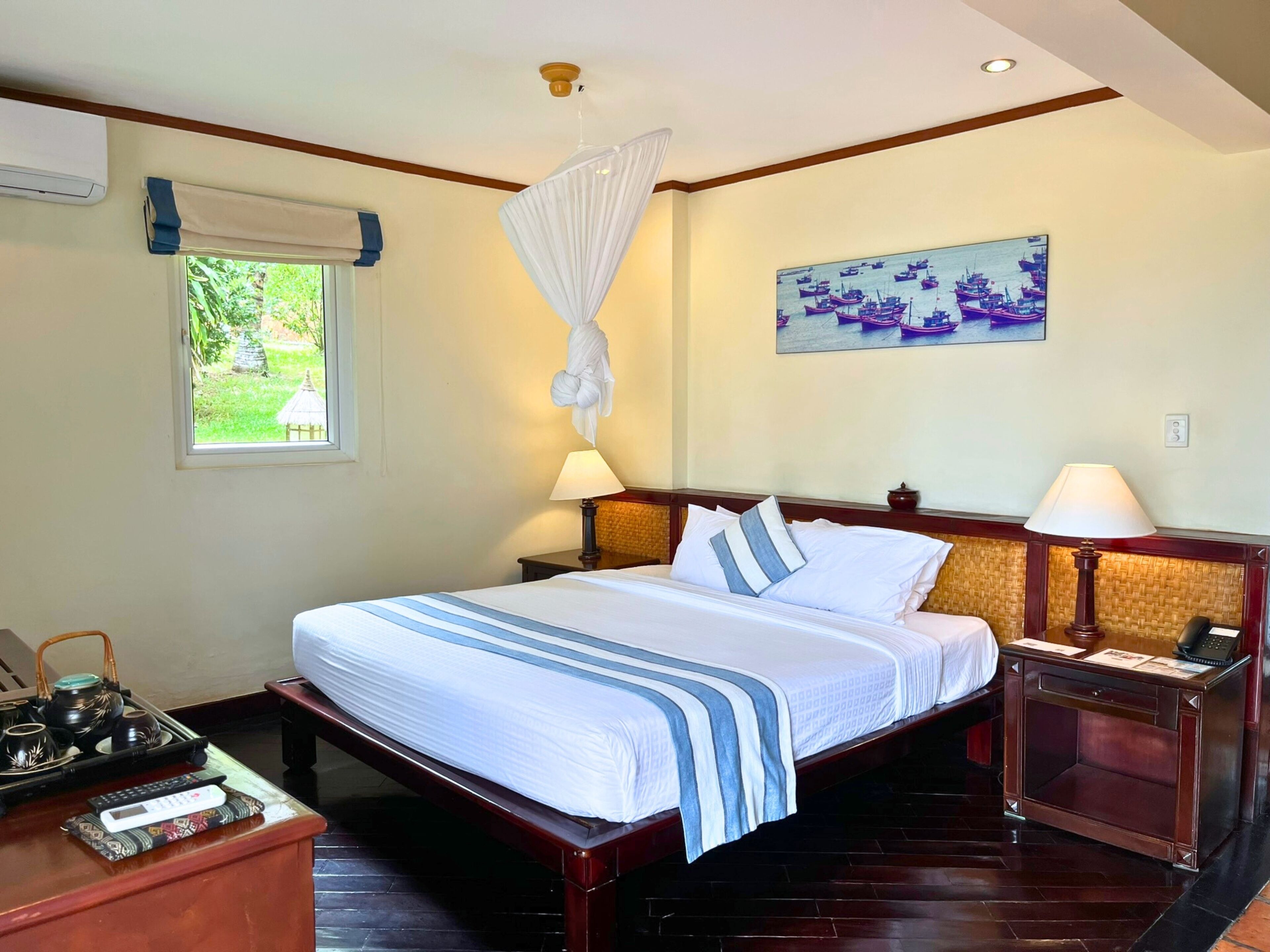 Una habitación de hotel con cama, mesitas de noche, un escritorio con teléfono, una lámpara, un cuadro en la pared y una ventana con vista a follaje.