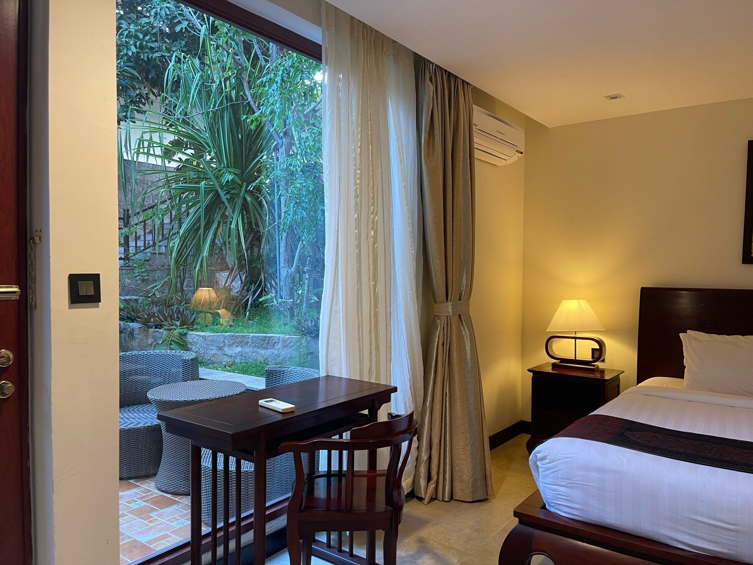 A hotel room with a bed, a desk, a chair, a nightstand with a lamp, and a view of greenery through a window.