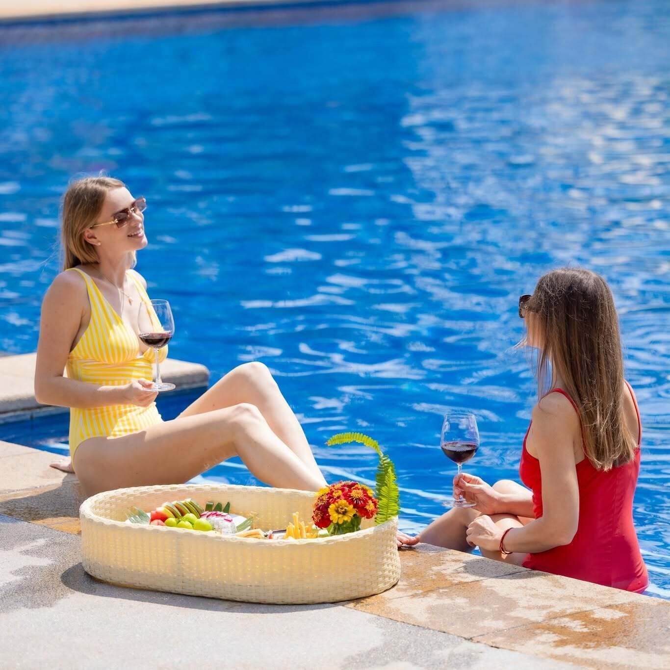 Dos personas sentadas junto a una piscina, con bebidas, una cesta de fruta y flores.