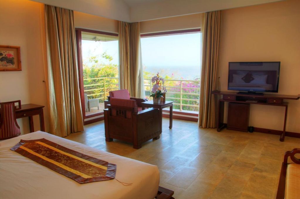 A hotel room with a bed, a desk with a television, a chair, and a balcony with curtains.