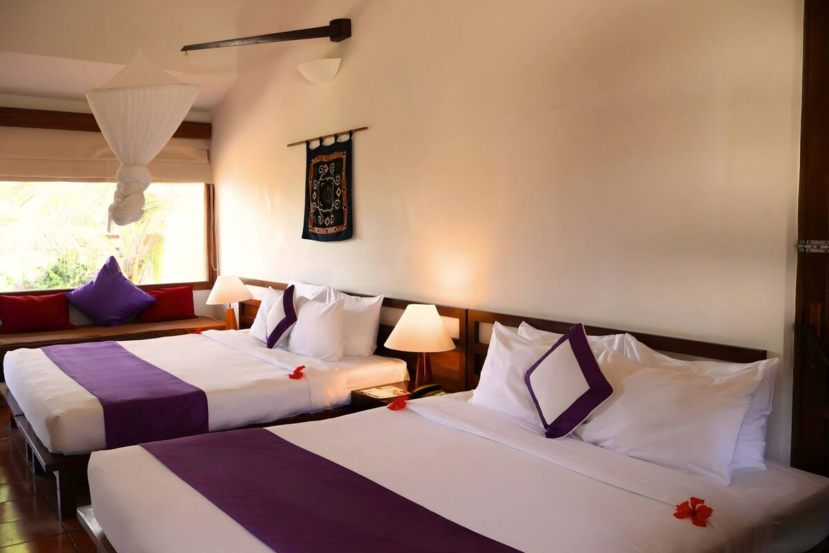 Une chambre d’hôtel avec deux lits, chacun agrémenté de draps blancs et de touches de couleur violette, une table de chevet avec une lampe et une décoration murale.