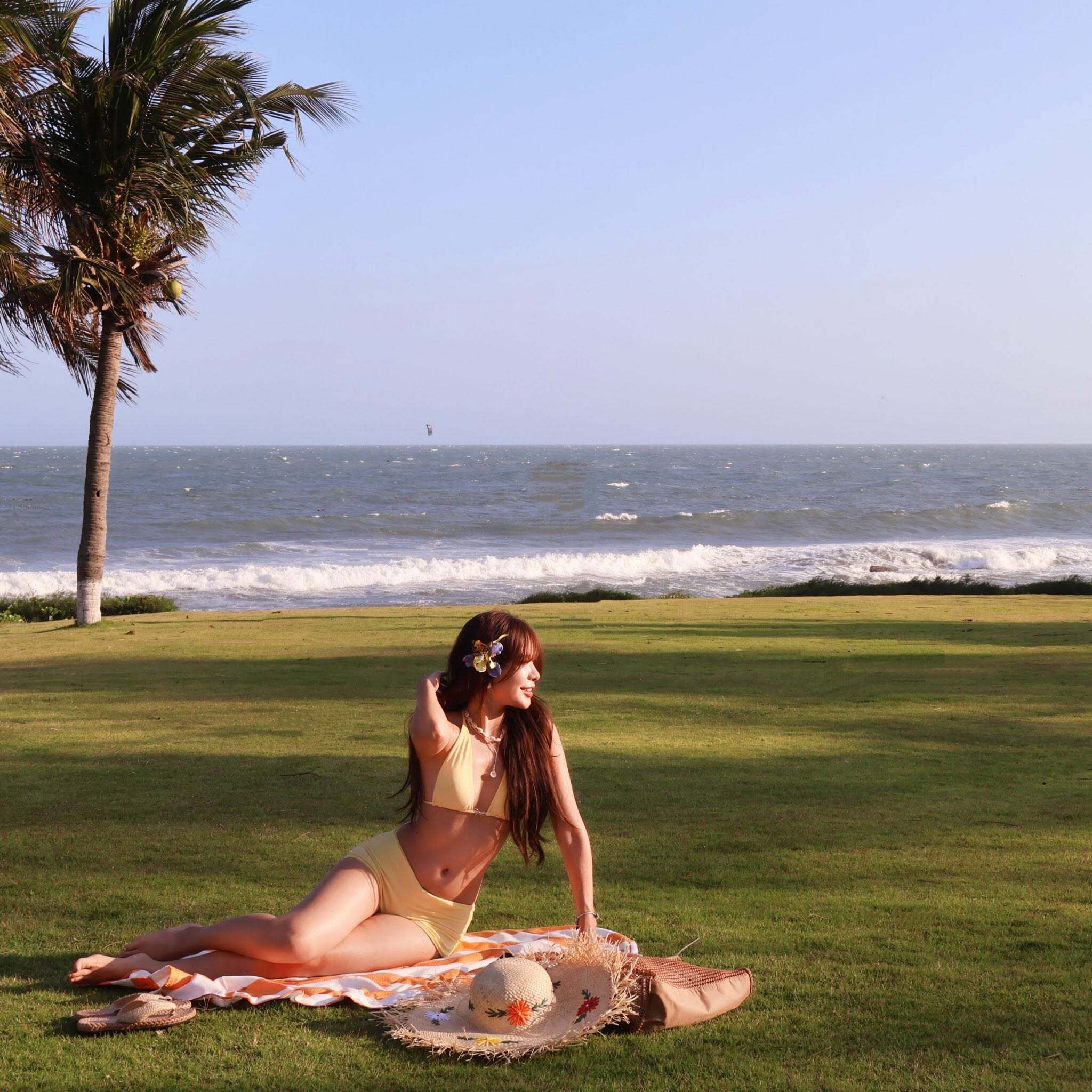 Una persona sentada sobre una manta en el césped, cerca de una palmera y el océano.